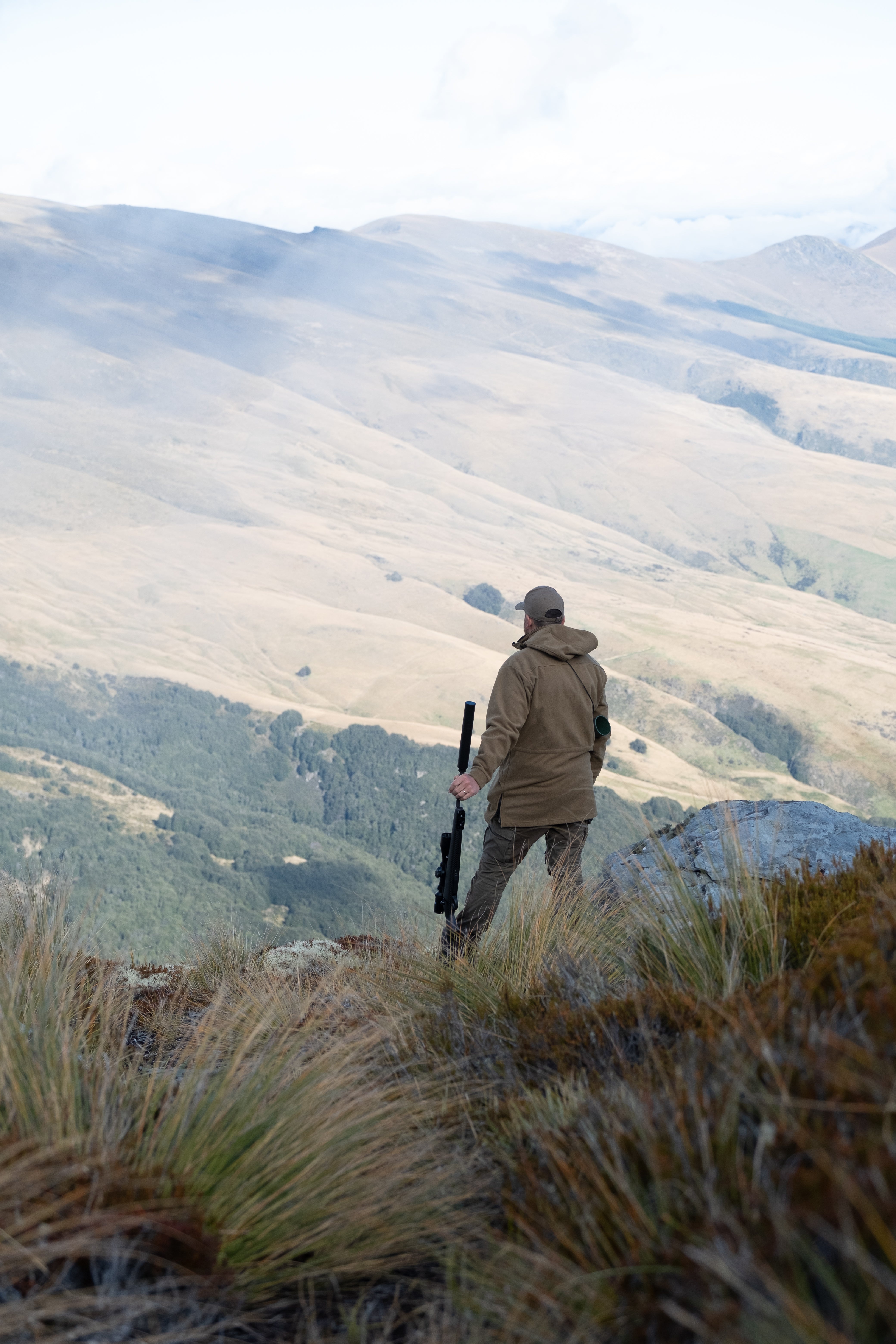 Person with a rifle standing on a mountain top with a scenic view of rolling hills.